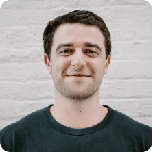 Smiling young man with short dark hair wearing a dark T-shirt standing in front of a white brick wall.